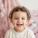 A happy toddler, aged 2-5, smiling directly at the camera. The background is softly blurred with dusty rose, white, and laven